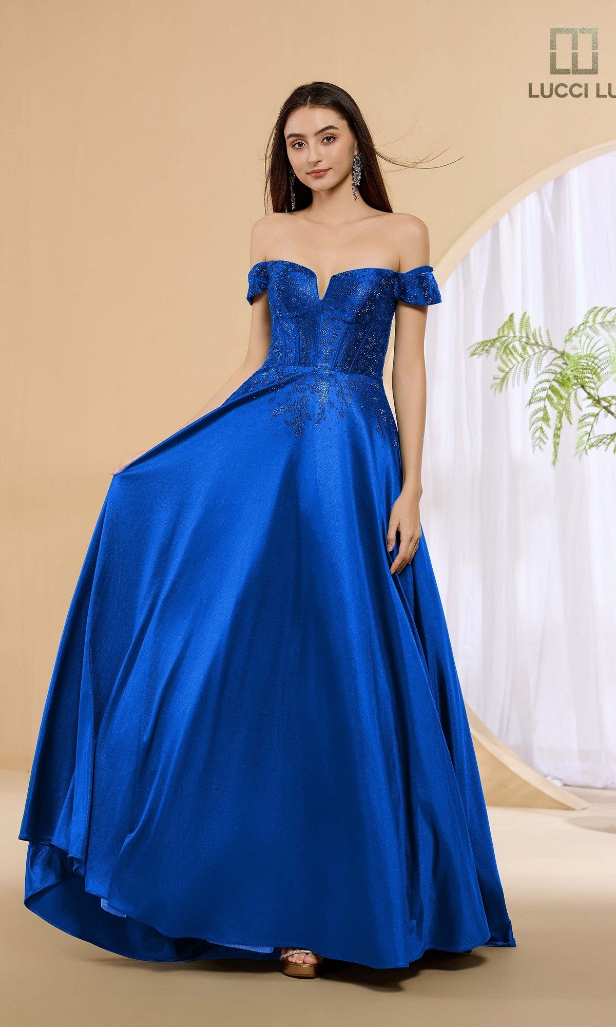 A woman poses in a studio wearing the Lucci Lu 1483 long royal prom dress, featuring an off-shoulder neckline, fitted corset bodice, and full a-line skirt made from luxurious stretch satin.